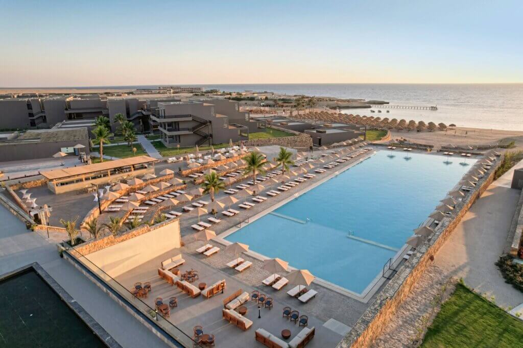 Ein großer, luxuriöser Swimmingpool im Freien, umgeben von Liegestühlen und Palmen, überblickt bei Sonnenuntergang den Sandstrand und das ruhige Meer in einem modernen Strandresort.