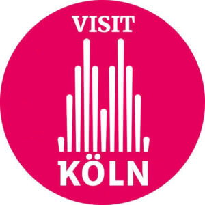 Ein roter Kreis mit der Aufschrift „VISIT KÖLN“ in weißer Schrift und einer stilisierten weißen Grafik, die dem Kölner Dom ähnelt.