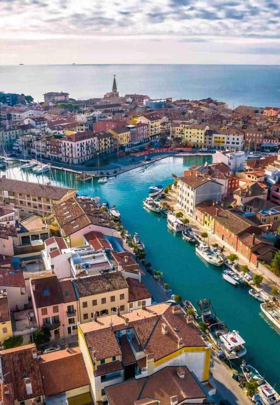 Luftaufnahme einer italienischen Küstenstadt mit farbenfrohen Gebäuden, engen, von Booten gesäumten Kanälen und einem entfernten Kirchturm, der sich unter einem teilweise bewölkten Himmel zum Meer hin öffnet.