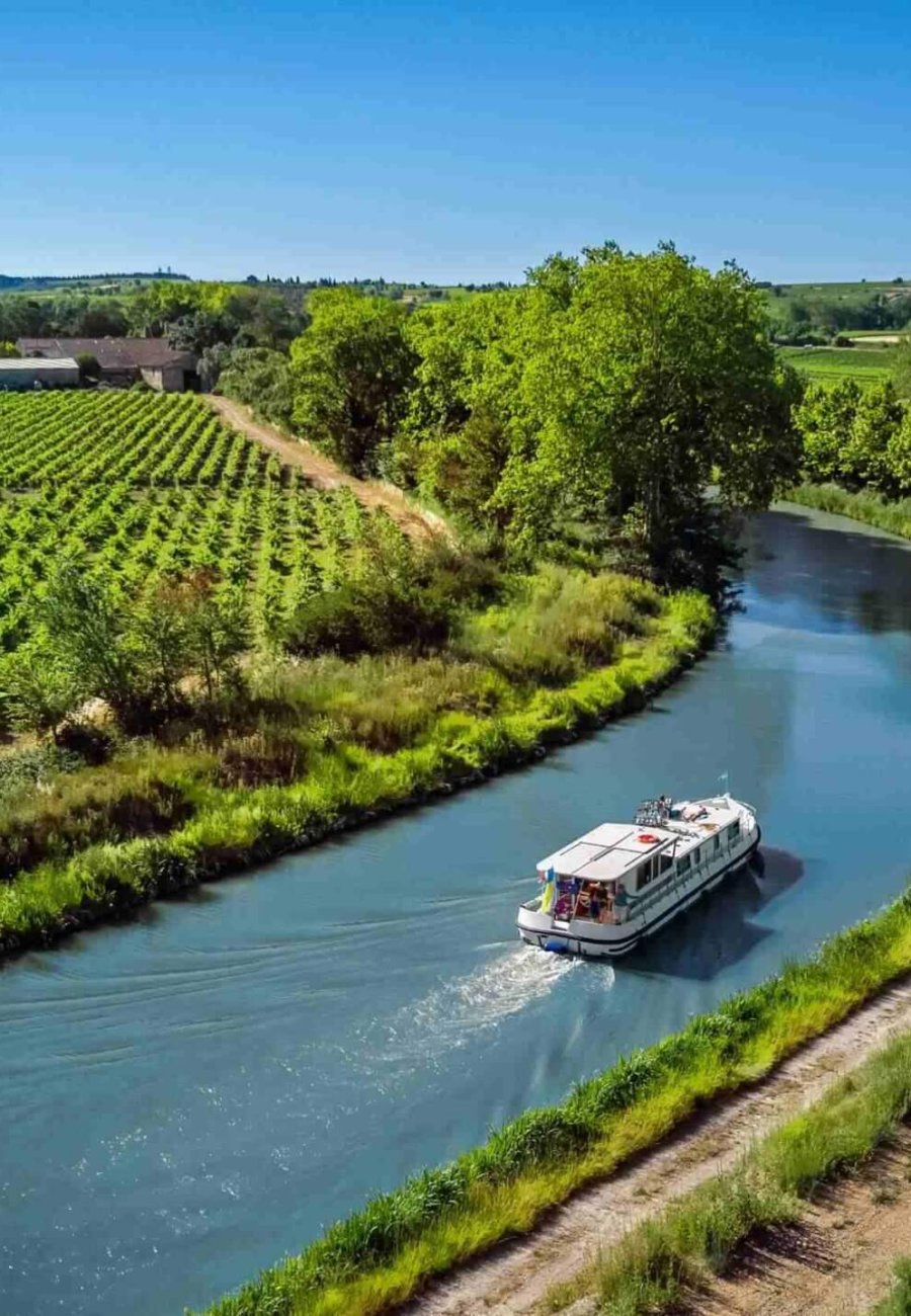 Ein Boot fährt auf einem gewundenen Kanal, der von Weinbergen und grünen Feldern gesäumt ist, unter einem klaren blauen Himmel, mit Bäumen und fernen Hügeln im Hintergrund.