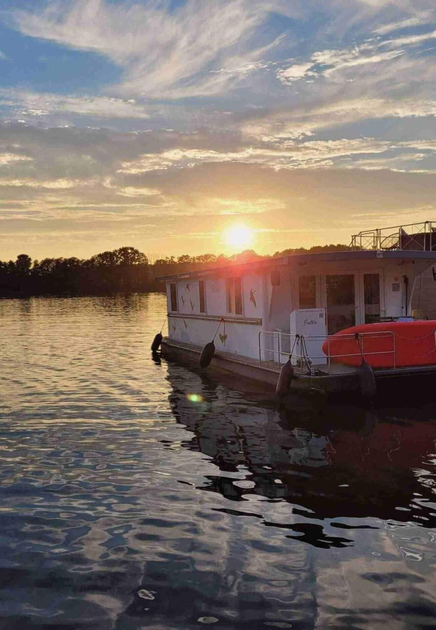 Ein Hausboot liegt bei Sonnenuntergang auf einem ruhigen See vor Anker. Das goldene Sonnenlicht spiegelt sich auf dem Wasser, und im Hintergrund zeichnen sich die Bäume gegen einen teilweise bewölkten Himmel ab.