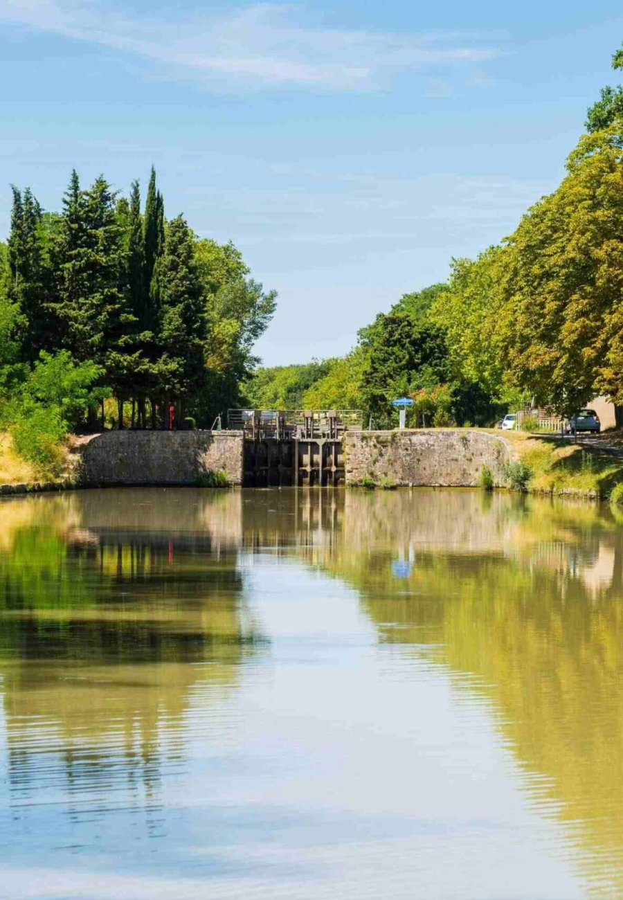Ein friedlicher, von grünen Bäumen gesäumter Kanal mit einem weißen Boot auf der rechten Seite und einer steinernen Schleusenbrücke im Hintergrund unter einem klaren blauen Himmel.